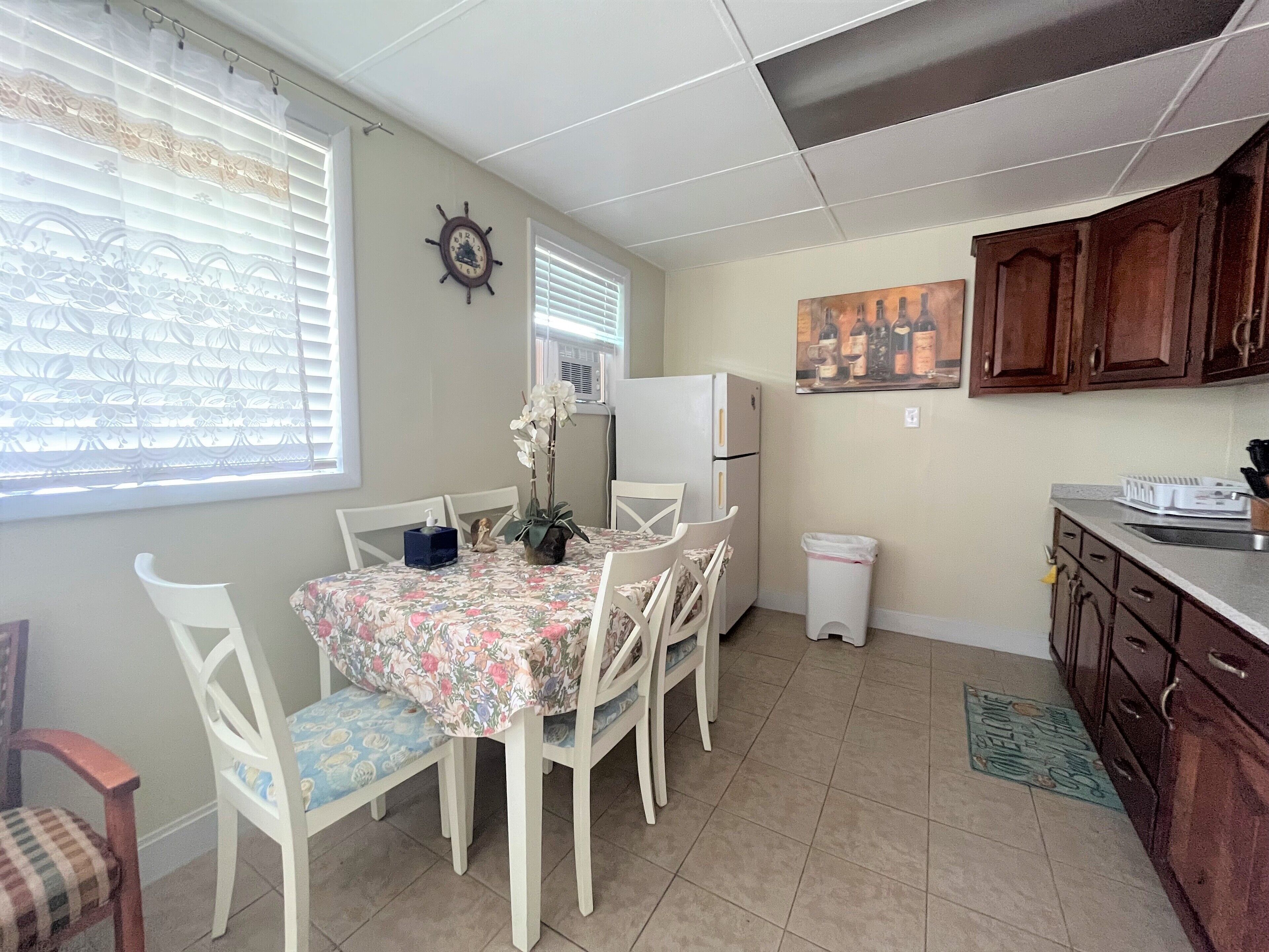 A dining area with a table set for four, white chairs, a refrigerator, and a clock on the wall.