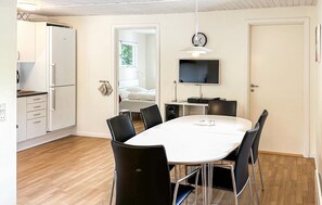 A modern kitchen with white cabinets, a built-in oven, and a refrigerator. There is a dining area with a round table and black chairs. A flat-screen TV is mounted on the wall above the table.