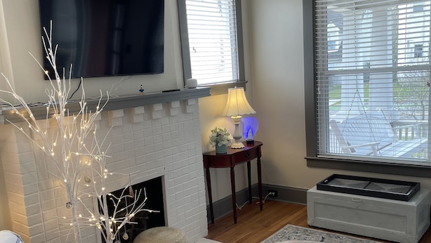 A living room with a fireplace, a flat-screen TV mounted above it, a large window with blinds, and a patterned rug.