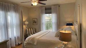 A bedroom with a bed, a ceiling fan, a window with curtains, a bedside table with a lamp, and a chair.