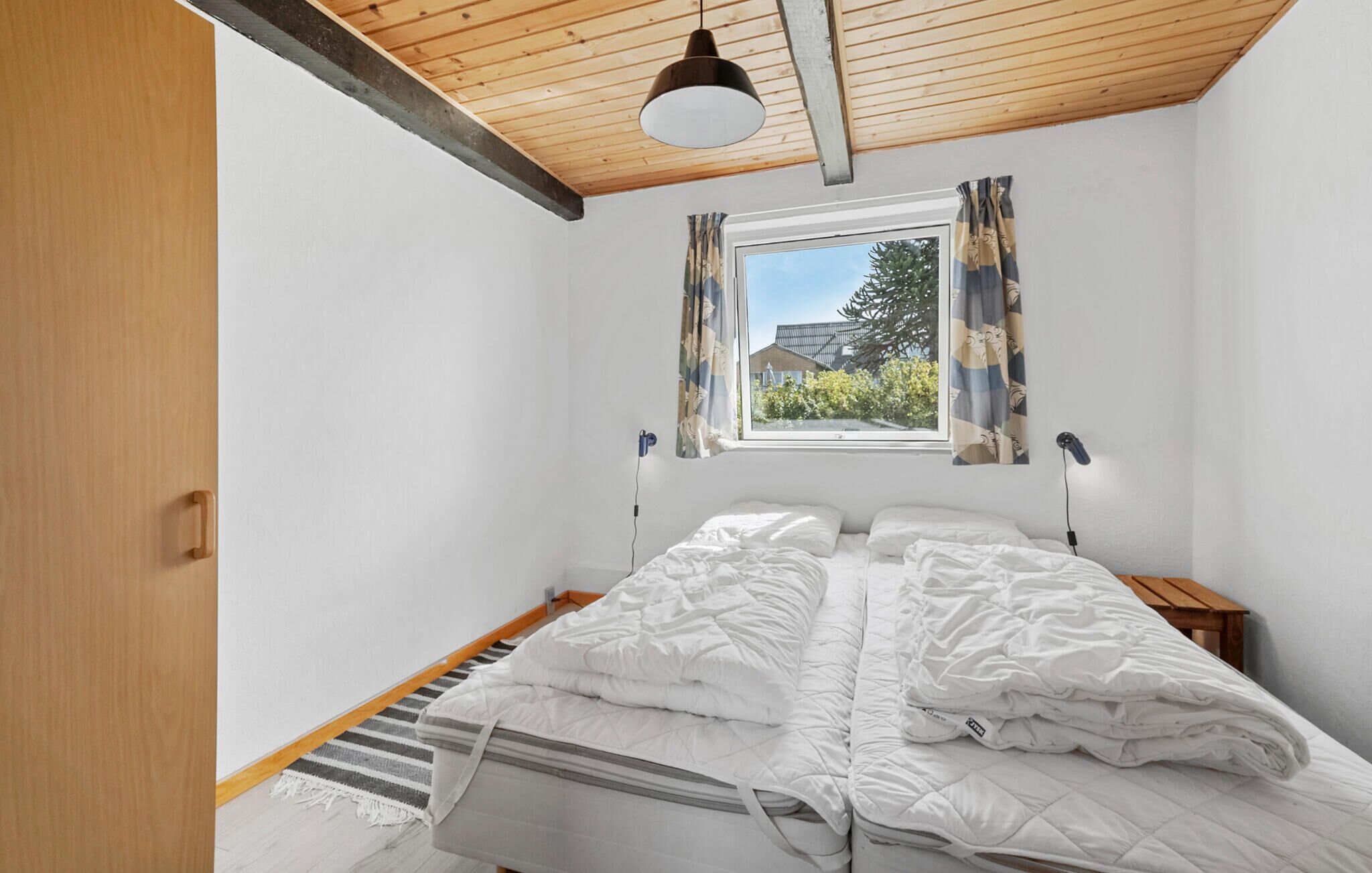 A bedroom with a wooden ceiling, a window with curtains, a bed with white bedding, and a bedside table.