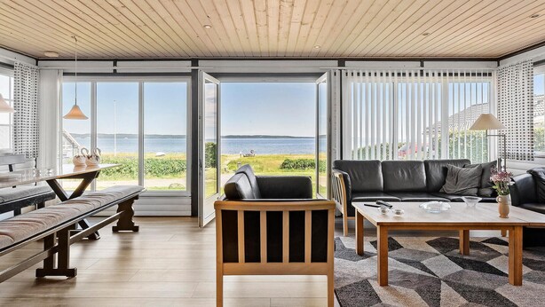 A spacious living area with a wooden ceiling, large windows offering a view of the sea, a black leather sofa, and a wooden dining table. - Lovely home in Fredericia with WiFi (Fredericia)