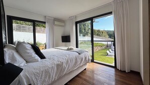 A modern bedroom with a large bed, a flat-screen TV, and a view of a garden through large windows.