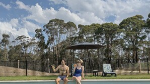Una piscina con dos personas sentadas en el borde, rodeada de césped y árboles.