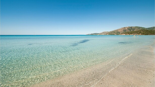 Una playa despejada con aguas poco profundas, una orilla de arena y un paisaje montañoso al fondo.
