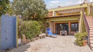 Un patio techado al aire libre con suelo de piedra, una silla azul, una mesa y un pequeño jardín con un árbol y plantas en macetas.