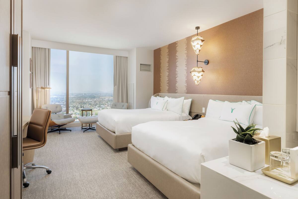 A hotel room with two beds, a desk, a chair, and a view of the city.