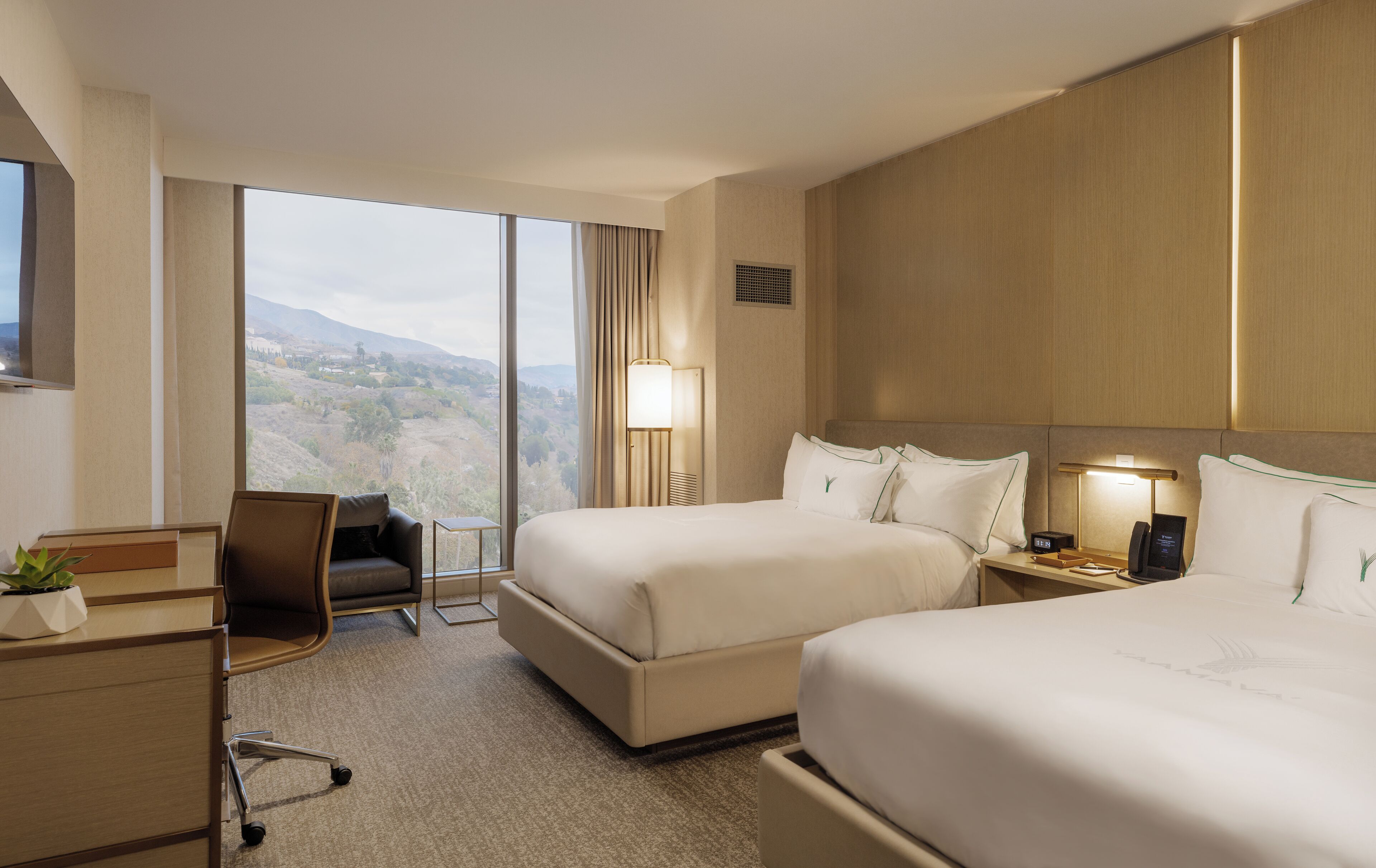 A hotel room with two beds, a desk, a chair, and a large window offering a view of the countryside.
