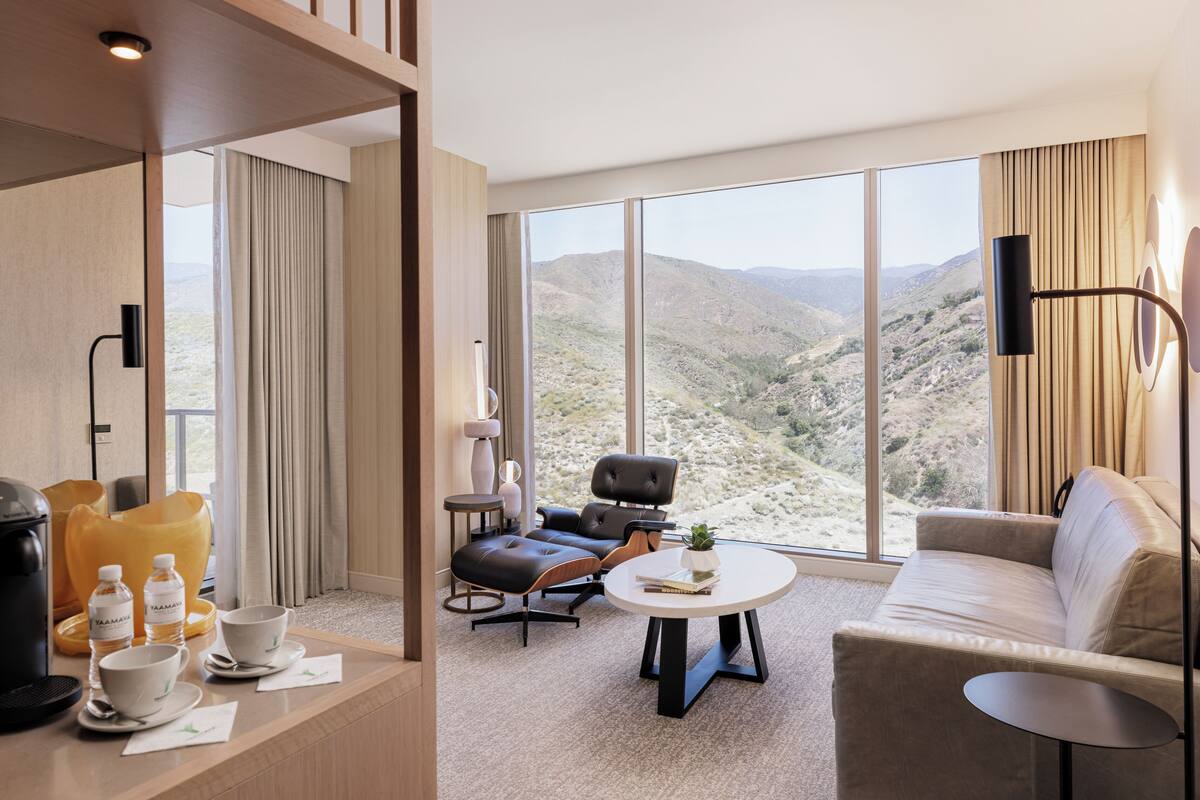 A modern hotel room with a large window offering a view of a mountainous landscape. There is a sofa, a chair, and a small table with a plant.