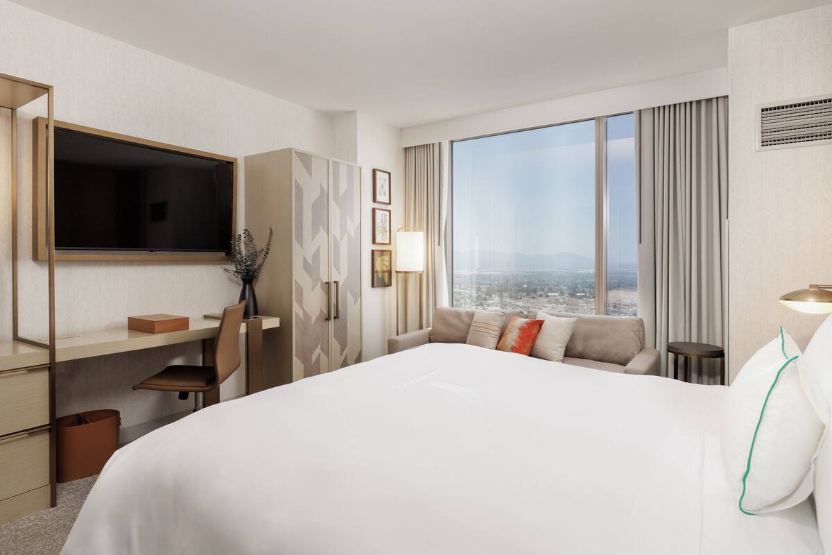 A hotel room with a bed, a desk, a TV, a city view, and a large window.