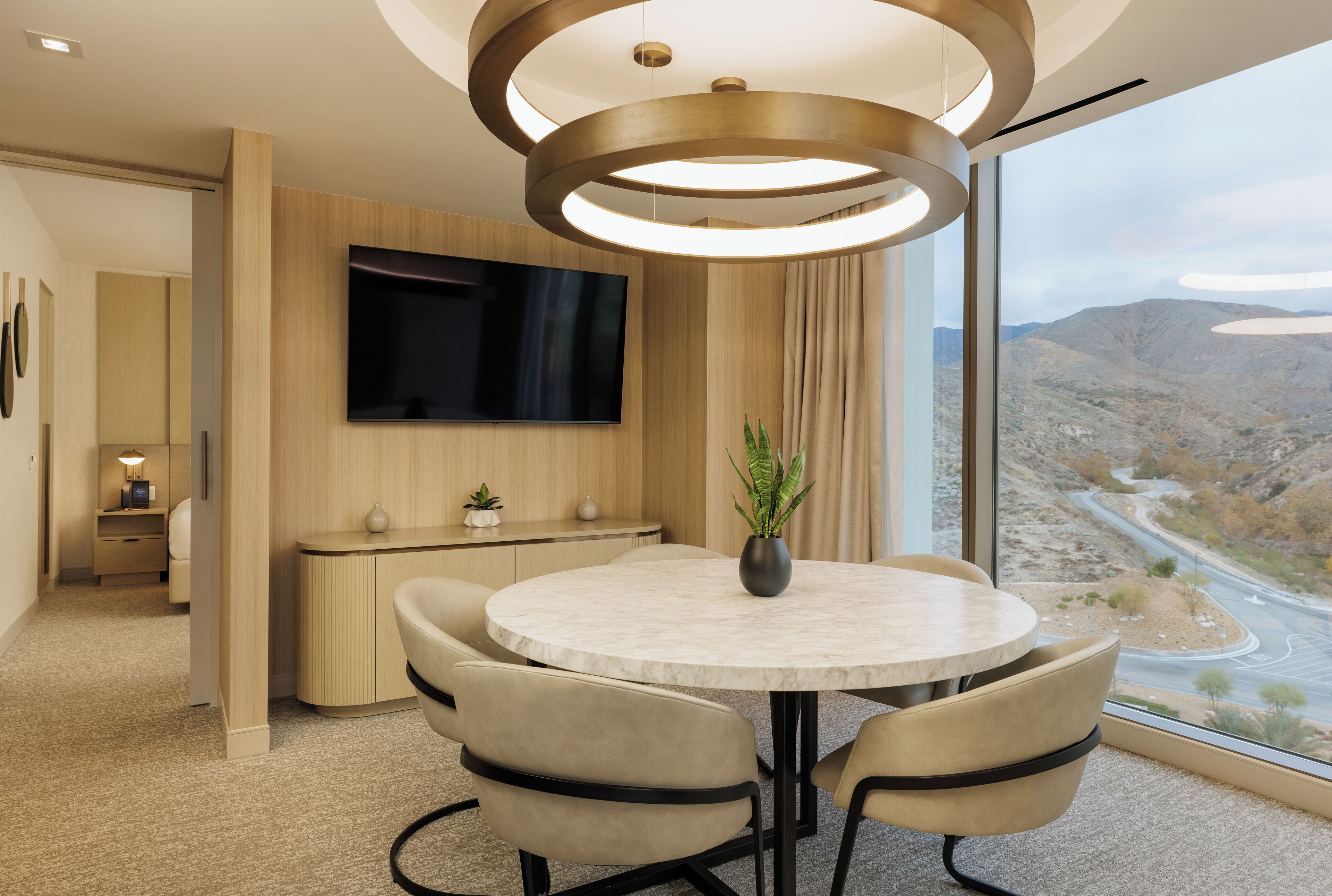 Una habitación de hotel moderna con una mesa de comedor redonda, asientos cómodos, un televisor de pantalla plana en la pared y un gran ventanal con vistas panorámicas de un paisaje montañoso.