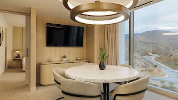 Una habitación de hotel moderna con una mesa de comedor redonda, asientos cómodos, un televisor de pantalla plana en la pared y un gran ventanal con vistas panorámicas de un paisaje montañoso.