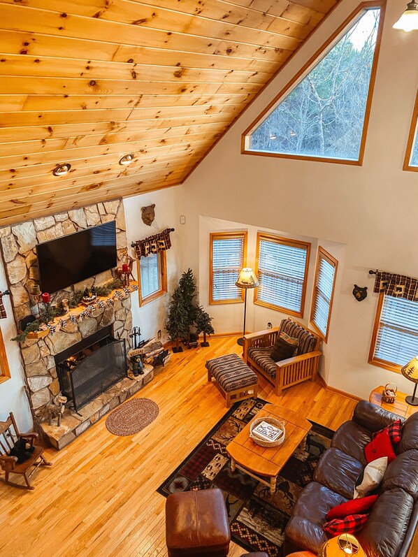 A spacious living room with a stone fireplace, wooden ceiling, and large windows.