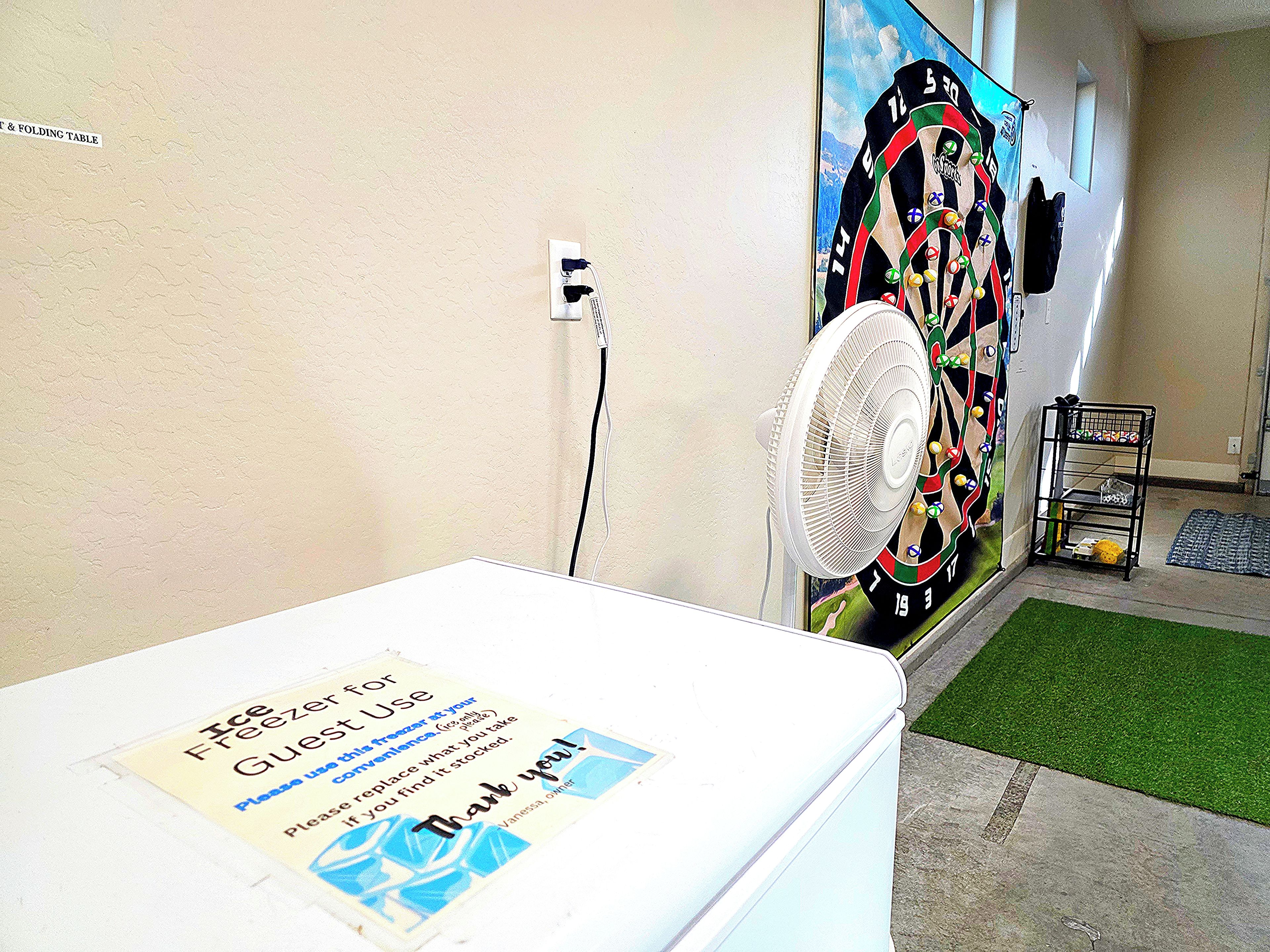 A room with a white table, a wall-mounted fan, a dartboard, and a small shelf.