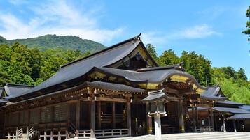 Un edificio japonés tradicional con techo curvo y estructura de madera, rodeado de un área de grava y una colina boscosa al fondo.