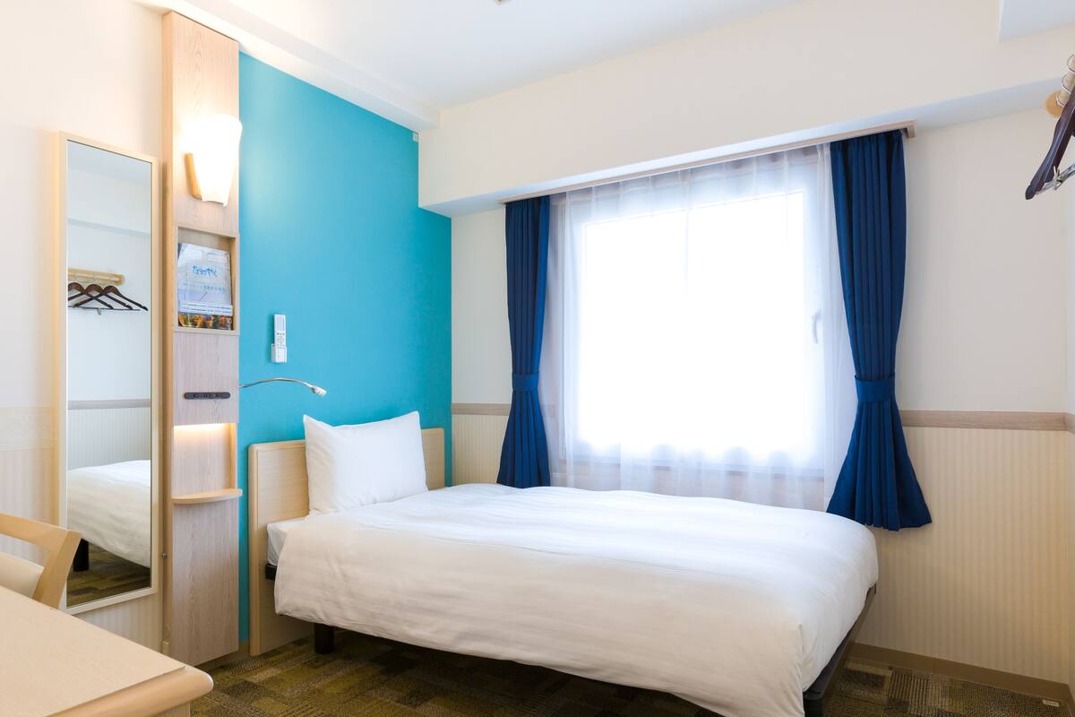 Une chambre d’hôtel avec un lit, un bureau, un miroir, une armoire et une fenêtre avec des rideaux.