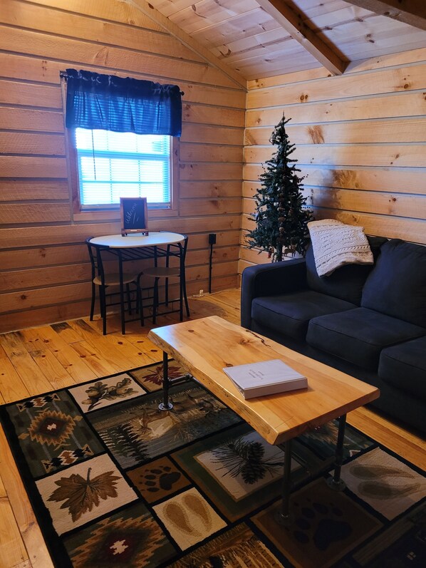A cozy cabin interior with a wooden table, a black sofa, a small Christmas tree, and a window with blinds.