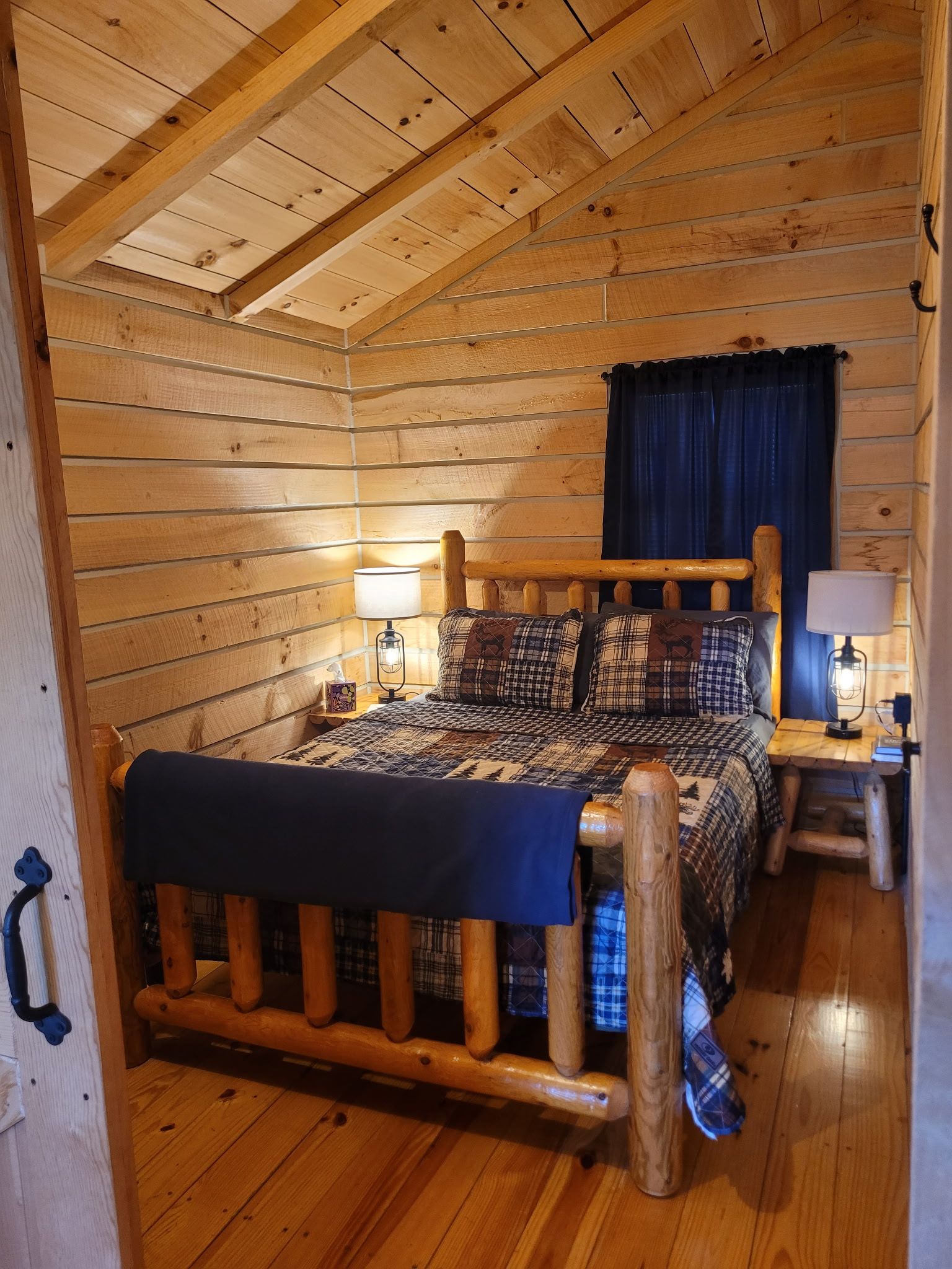 A log cabin bedroom with a wooden bed, two bedside lamps, a blue curtain, and a wooden headboard.
