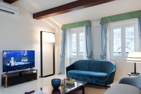 A modern living room with a blue sofa, a flat-screen TV, a floor lamp, and a window with curtains.