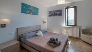 Ein Schlafzimmer mit Bett, Nachttisch, Lampe, Fenster mit Blick und einem Bild an der Wand.