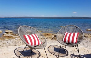 Zwei Korbstühle mit roten und weißen Streifenpolstern auf einer felsigen Küste mit Blick auf ein ruhiges Meer und ferne Hügel.