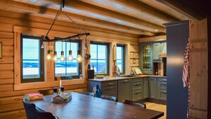 Interior de una cabaña de madera con comedor, cocina y ventanas grandes que ofrecen vistas panorámicas.
