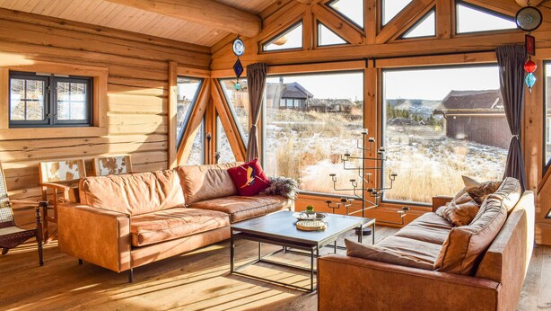 Interior acogedor de una cabaña con paredes de madera, un sofá, una mesa de centro y grandes ventanales que ofrecen vista al paisaje nevado.