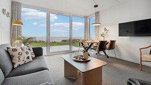 Een moderne woonkamer met een groot raam met uitzicht op zee, een bank met marine-blauwe kussens, een eethoek met stoelen, en een flatscreen televisie aan de wand.