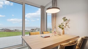 Een eethoek met uitzicht op zee, een tafel met boeken en een fles, en een vaas met groene planten.