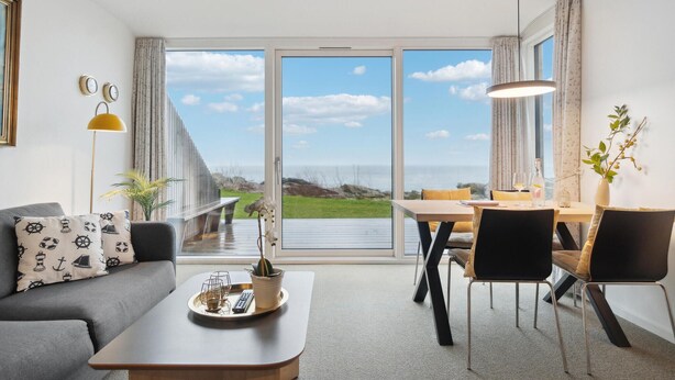 Een moderne woonkamer met een bank, een salontafel, en een eethoek met stoelen en een tafel. Er is een groot raam met uitzicht op de zee.