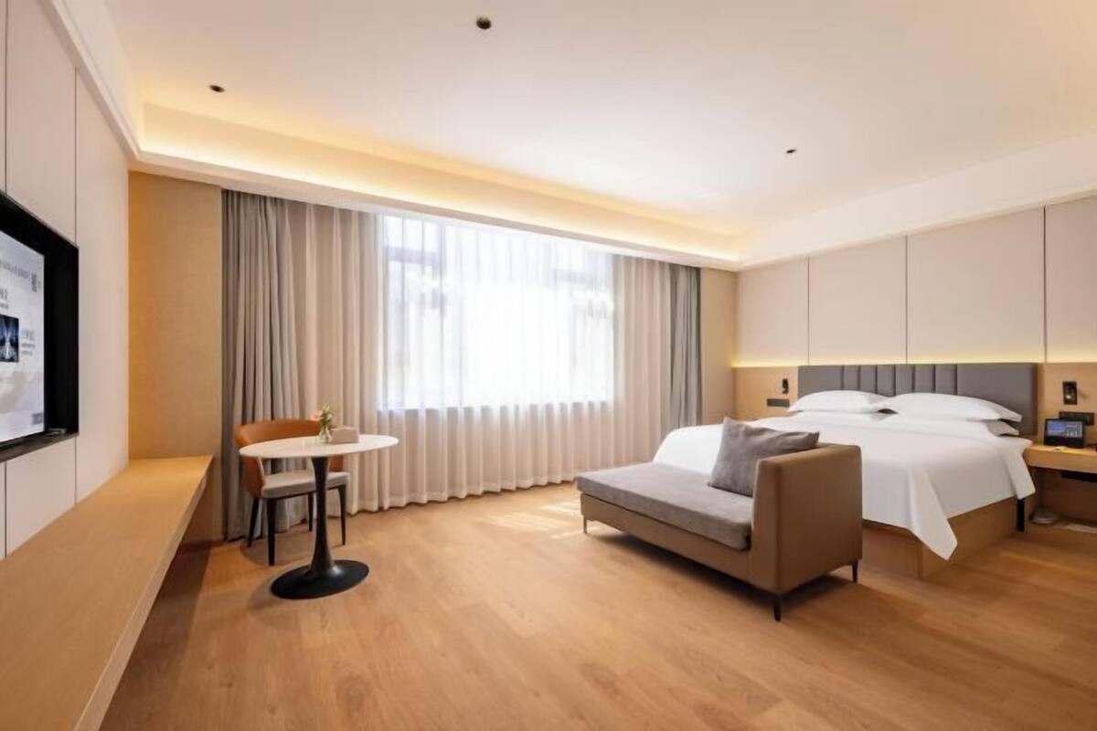 Habitación de hotel con cama, sofá, una mesita, televisor y un ventanal con cortinas.