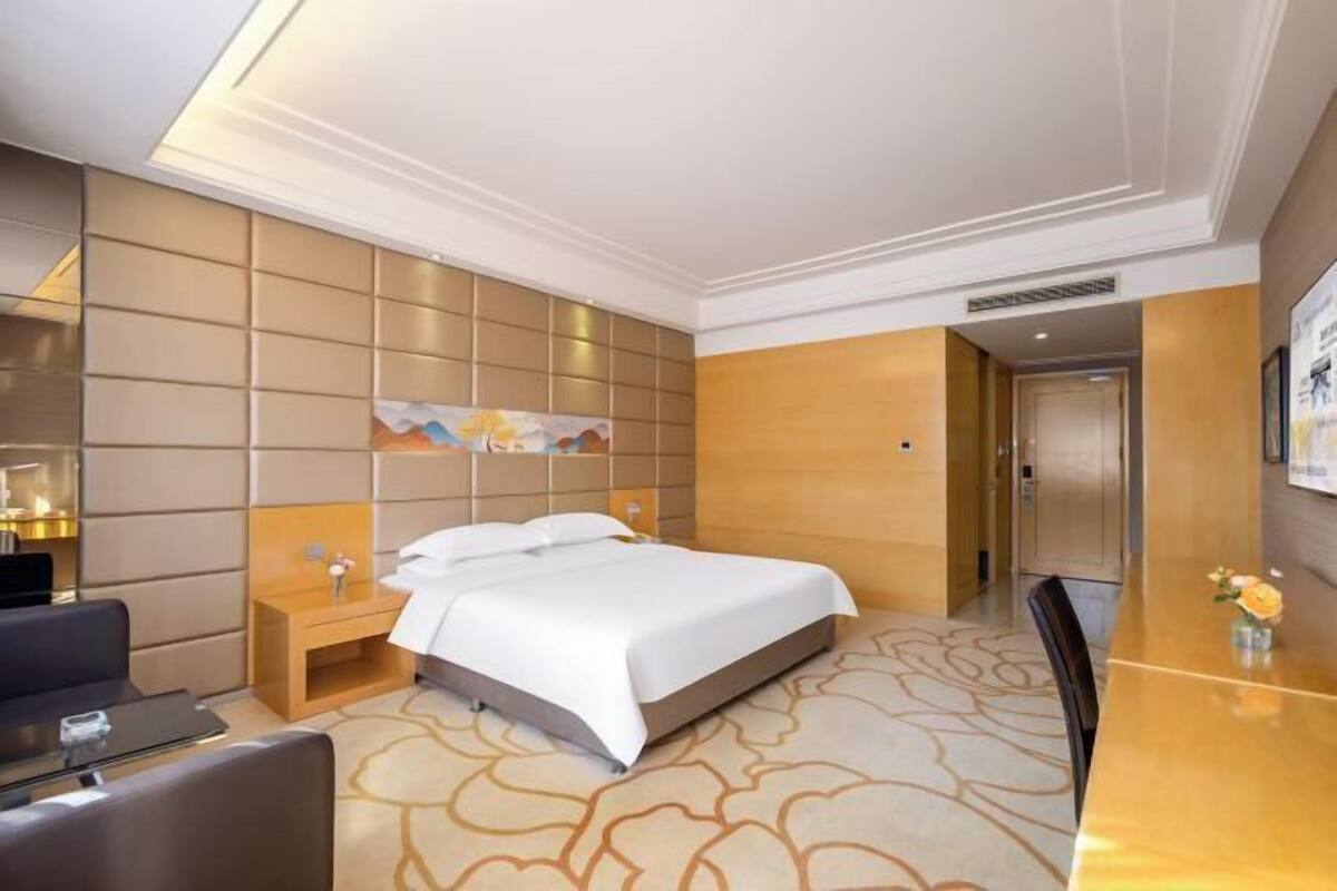Una habitación de hotel con cama, mesita de noche, silla y escritorio con un jarrón de flores.