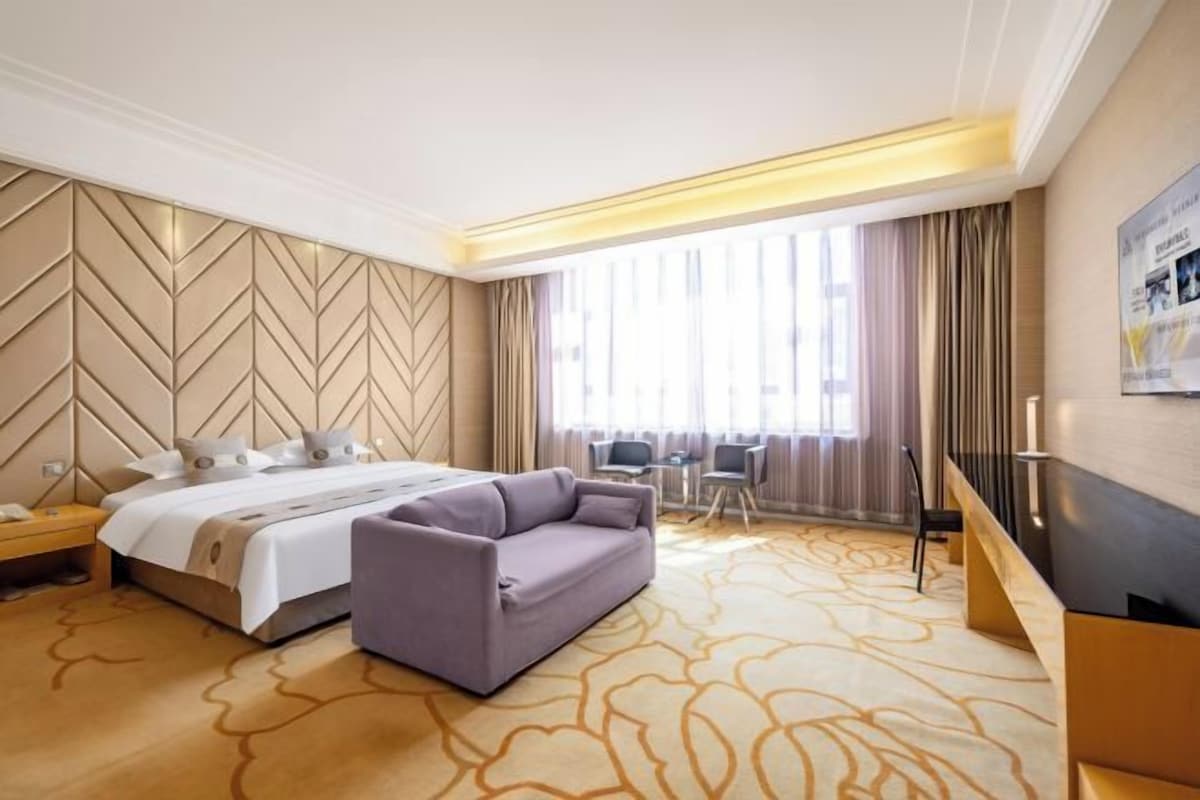 A hotel room with a bed, a sofa, a desk, and a large window with curtains.