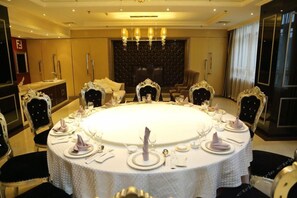 Une table de salle à manger ronde, mise pour un repas, avec des chaises, un lustre et une grande fenêtre avec des rideaux.