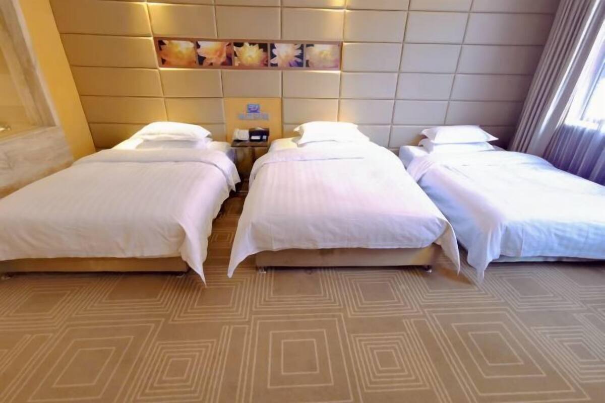 Habitación de hotel con tres camas individuales, cabecera decorada con motivos florales, mesita de noche con lámpara y alfombra estampada.