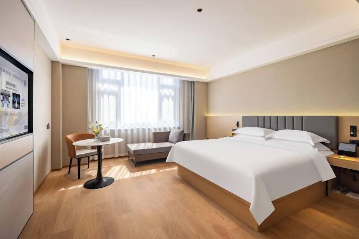 Una habitación de hotel moderna con una cama grande, una mesita redonda pequeña con una silla, un sofá y un ventanal con cortinas.