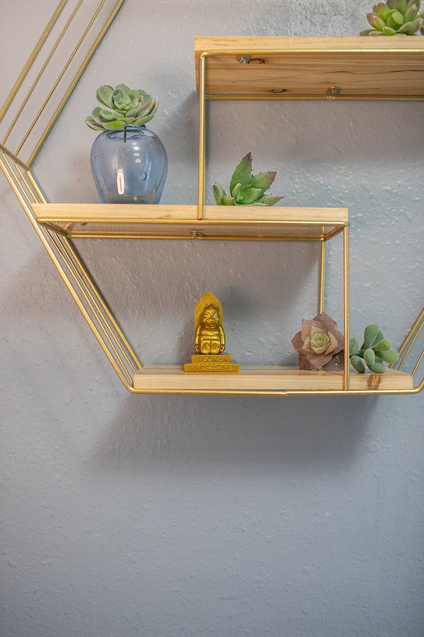 A geometric wall shelf with a golden frame, holding a small blue vase with succulent plants, a golden Buddha statue, and a pink flower.