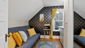 A cozy attic bedroom with a dark patterned wall, a small window with yellow curtains, a microwave on a wooden stand, and a blue sofa with patterned cushions. - Billiken Inn, Private Loft 2nd floor Loft in 2 Story Victorian  (Spokane)