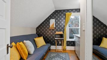 A cozy attic bedroom with a dark patterned wall, a small window with yellow curtains, a microwave on a wooden stand, and a blue sofa with patterned cushions.