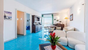 A spacious living room with a blue-tiled floor, a white sofa set, a dining area with a table and chairs, and a view through a glass door. - Cozy apartment in Ischia with WiFi (Ischia)