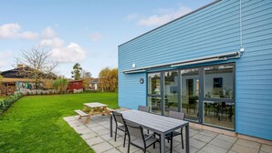 A patio with a table and chairs, a blue building with large windows, and a grassy lawn.