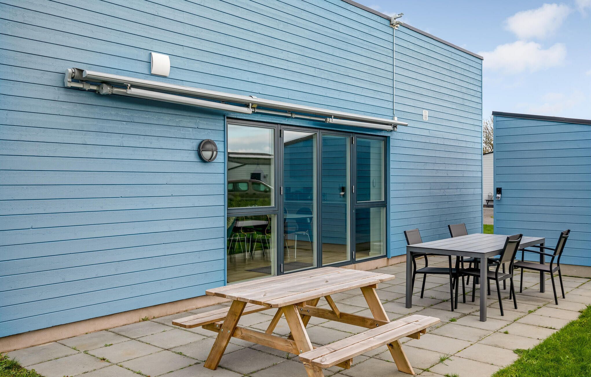 Un patio con una mesa y sillas de picnic de madera, una puerta de cristal y fachada de edificio azul.