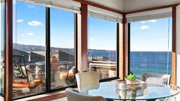 A balcony with a glass table and chairs, overlooking a body of water and a distant shoreline. - Coastal Luxury: Oceanfront Views, Decks + Walkable to Downtown (Monterey)