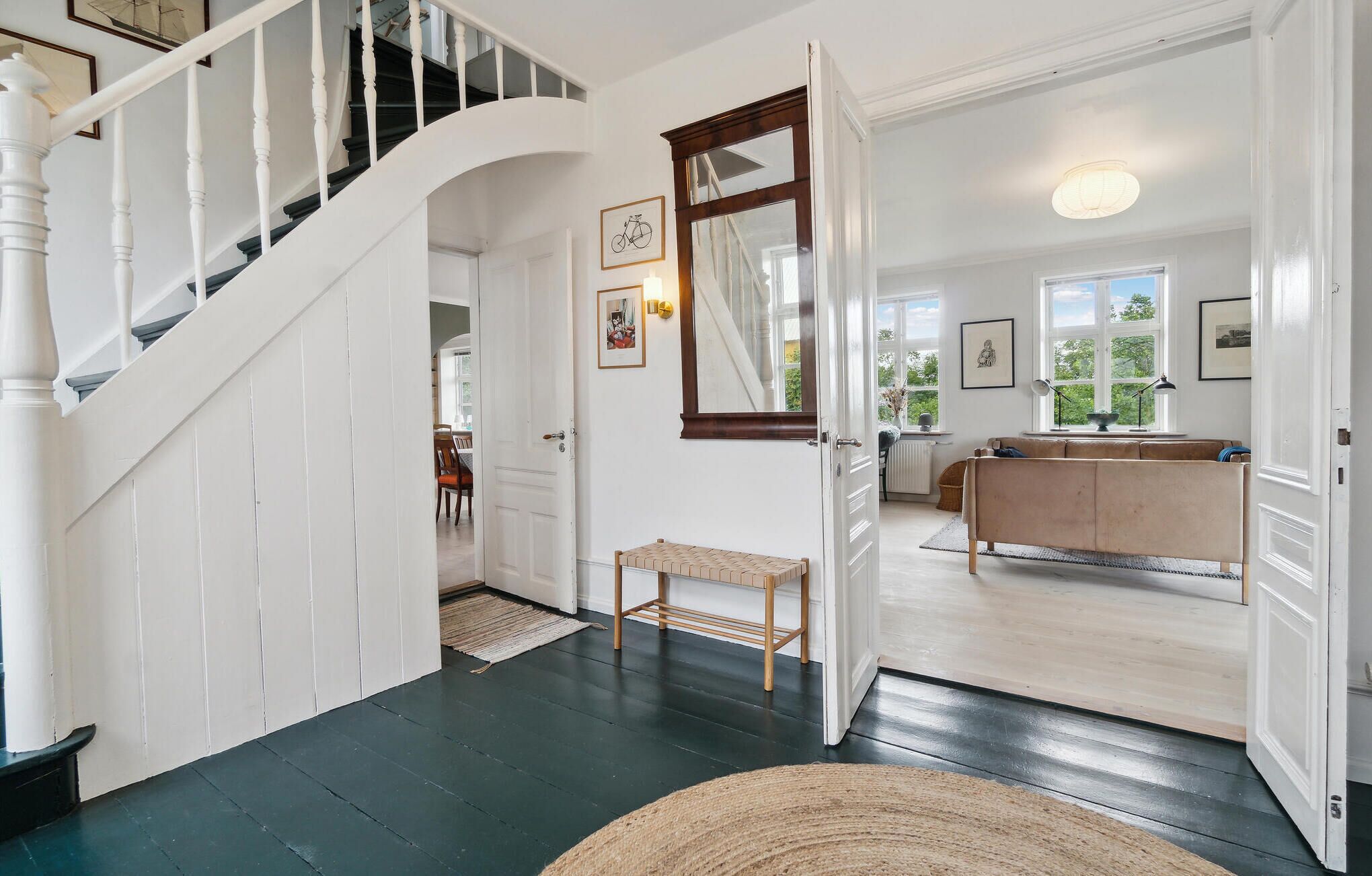 A spacious hallway with a dark wooden floor, a white staircase, and a dining area visible through an open door.
