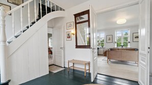 Un hall d’entrée spacieux, doté d’un parquet en bois foncé, d’un escalier blanc et d’un coin repas visible à travers une porte ouverte.