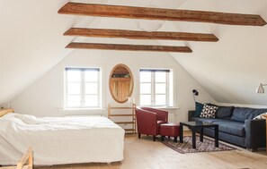 Une chambre mansardée chaleureuse, avec un lit, un fauteuil rouge, une table noire et un canapé bleu. Il y a deux fenêtres et une poutre en bois au plafond.