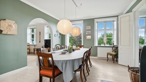 Une salle à manger avec une grande table, des chaises et des lanternes en papier suspendues au plafond. De grandes fenêtres offrent une vue sur la verdure environnante.