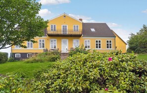 Une maison jaune à deux étages, dotée d’un balcon, entourée de verdure et d’une pelouse bien entretenue.
