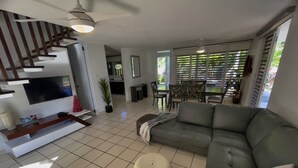 Una sala de estar moderna con un sofá gris, un televisor sobre un mueble blanco, un área de comedor con una mesa y sillas, y una escalera con barandilla de madera.