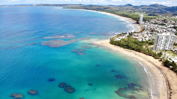 Vista aérea de una zona costera con aguas turquesas cristalinas, una playa de arena y una área residencial con edificios y vegetación.